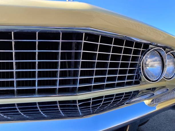 Classic car's front grille and polish chrome bumper highlighted by a vibrant blue sky emphasizing its timeless beauty and craftsmanship