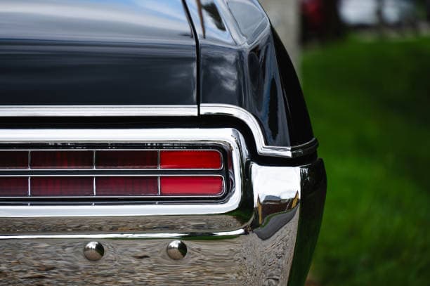 The tail lights of a classic car featuring a retro style with bright red lenses and a polish chrome bumper highlighting its timeless elegance