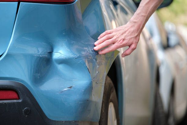 A person examines the front of a damaged car, assessing the extent of the damage with a concerned expression while considering the bumper repair and paint cost