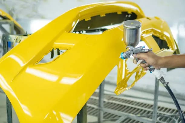 A person meticulously painting a yellow car with a spray gun highlighting the vibrant color and craftsmanship while also considering a quick fix for bumper falling