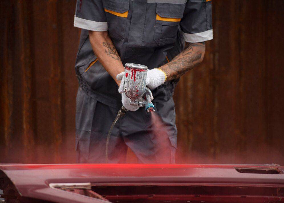 A worker in uniform meticulously sprays red paint onto a car, demonstrating professional painting techniques while considering the overall bumper repair and paint cost